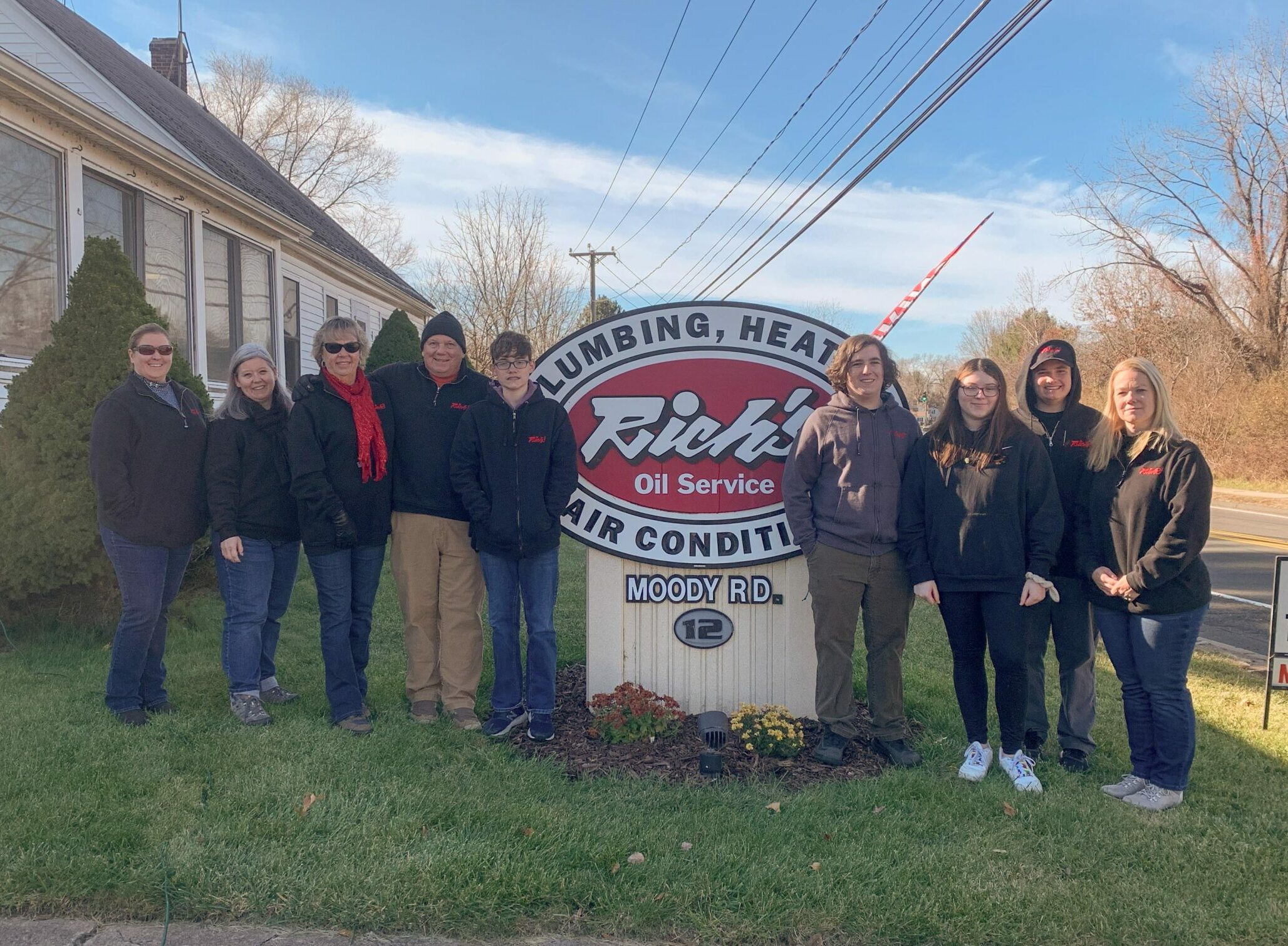 Family of 9 standing around a sign that says "rich's oil service" there is blue sky and winter weather